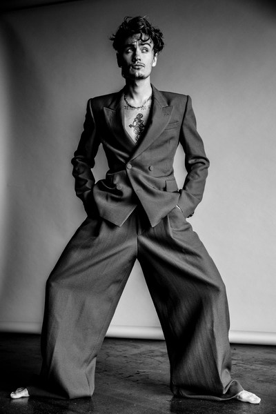 Young adult man in a stylish suit poses in a black and white studio shot, hands in pockets, looking slightly away from the camera.