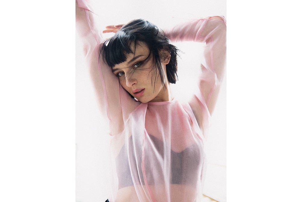 Portrait of a young woman with short dark hair and pink top, posing with her arms raised against a bright white background.