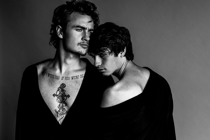 Monochrome portrait of two young adult men with dark hair in a close embrace, wearing dark shirts against a plain grey background.