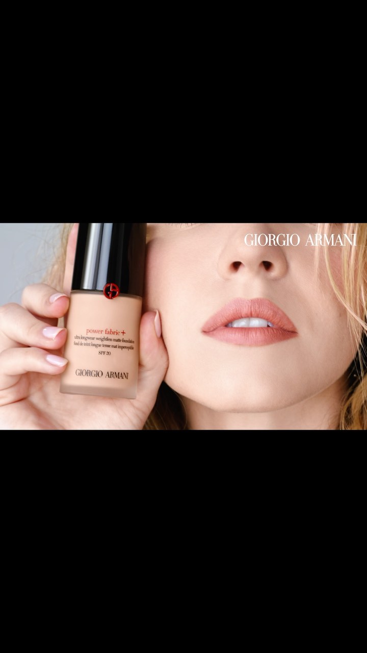 Close-up of a fair-skinned woman holding Giorgio Armani foundation; beige bottle with black cap, pink lips, and blond hair are visible.