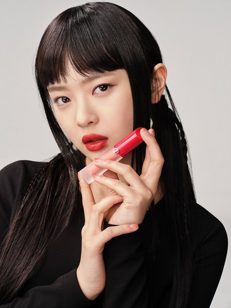 Asian woman with red lipstick holds a tube of lipstick in a close-up shot on a light background for a beauty advertisement.