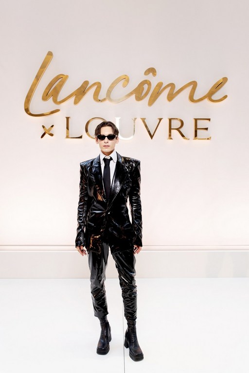 Fashionable adult in glossy black suit, tie, and sunglasses posing against a branded wall with Lancôme and Louvre logos in gold.