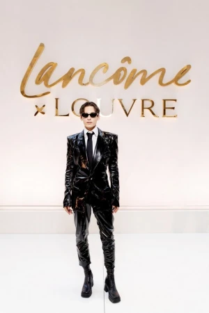 Fashionable adult in glossy black suit, tie, and sunglasses posing against a branded wall with Lancôme and Louvre logos in gold.