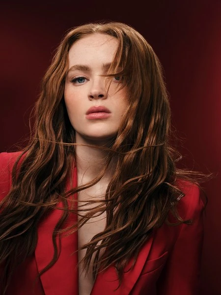 Portrait of a young woman with auburn hair wearing a red blazer against a dark red background in a head and shoulders shot.