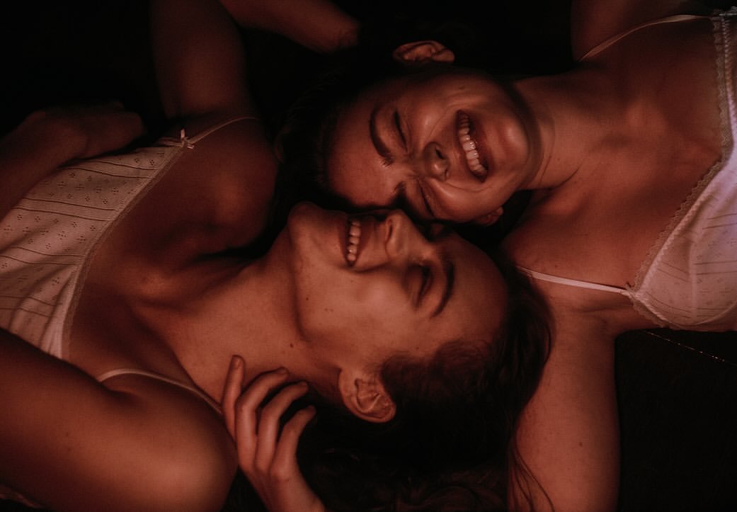 Two adult females, close together, laughing in low light indoors, wearing white tops. Portrait captures joy and intimacy.