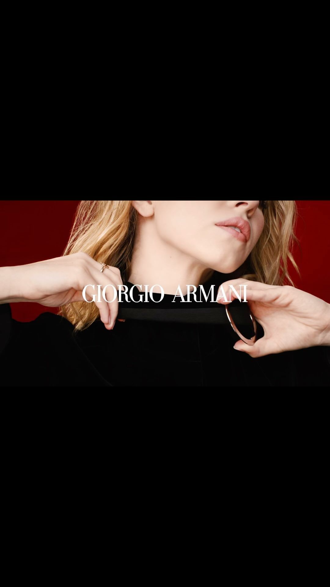 Close-up of a blonde woman in black, holding a black and gold beauty tool against a red backdrop, featuring Giorgio Armani branding.