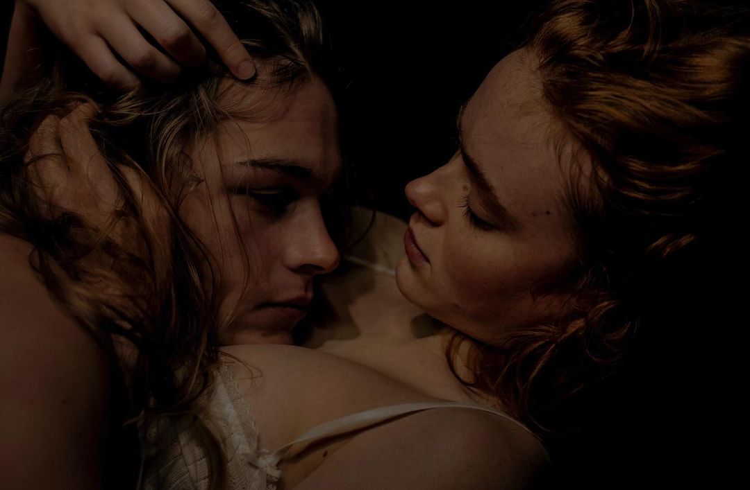 Close-up shot of two young women intimately posed in dark setting, one with auburn hair, the other with blonde hair. Intimate portrait.