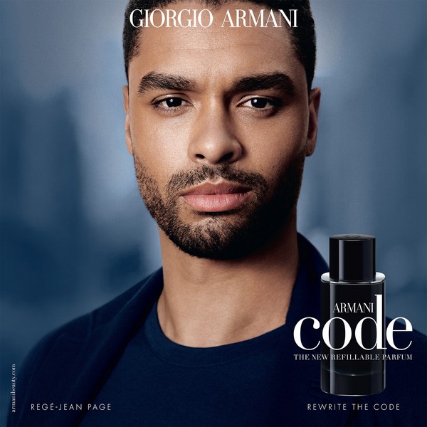 Close-up studio shot of a black male model with stubble wearing a dark navy jacket. The image also features a bottle of Armani Code cologne.