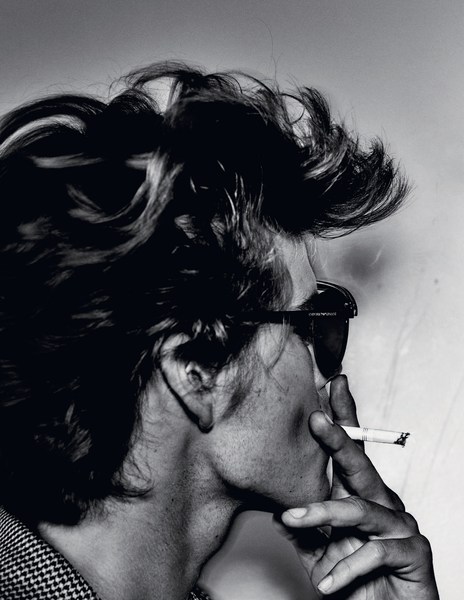 Black and white portrait of an adult with tousled hair and sunglasses smoking a cigarette against a plain backdrop, showcasing editorial style.