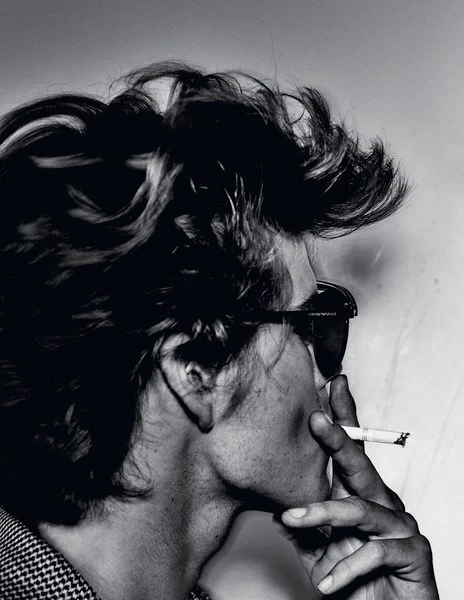 Black and white portrait of an adult with tousled hair and sunglasses smoking a cigarette against a plain backdrop, showcasing editorial style.