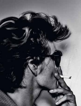 Black and white portrait of an adult with tousled hair and sunglasses smoking a cigarette against a plain backdrop, showcasing editorial style.