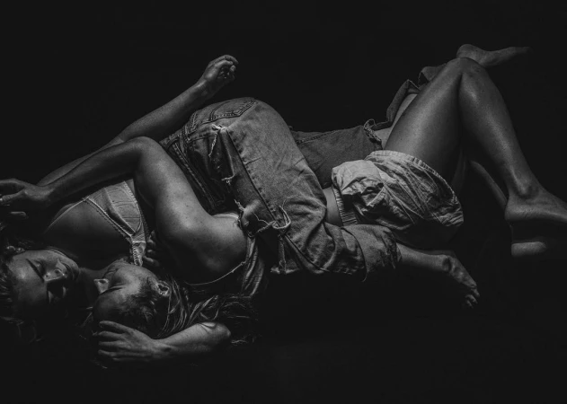 Monochrome photographic portrait of two people embracing closely while lying down against a dark background.