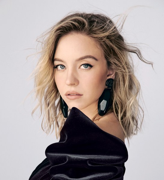 Close-up portrait of a blonde woman with blue eyes wearing a black dress and large earrings against a white background in studio lighting.