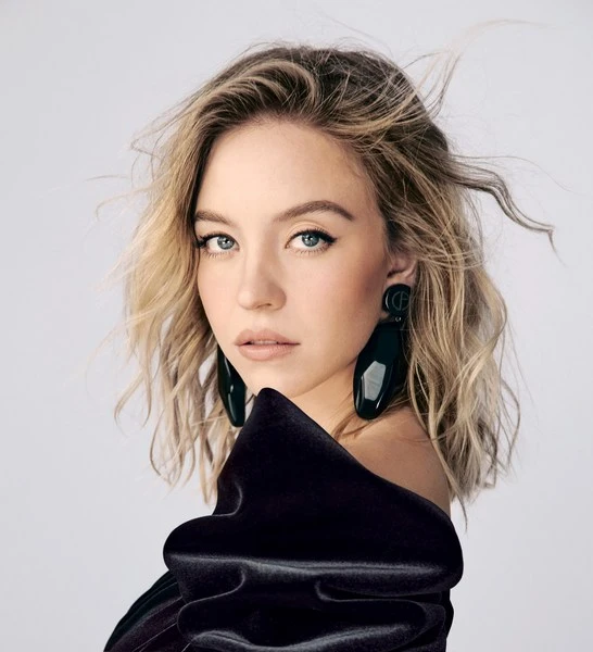 Close-up portrait of a blonde woman with blue eyes wearing a black dress and large earrings against a white background in studio lighting.