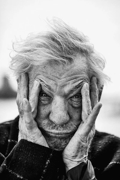 Monochrome close-up portrait of an elderly man with white hair, his hands framing his face, set against an out-of-focus background.