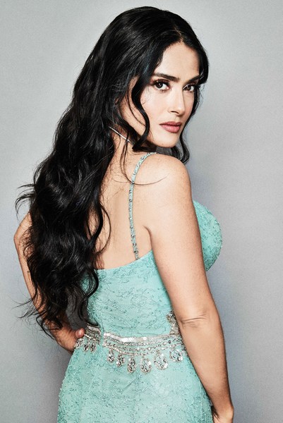 Salma Hayek poses in a light blue dress, with long dark hair, looking over her shoulder in a portrait against a gray backdrop.
