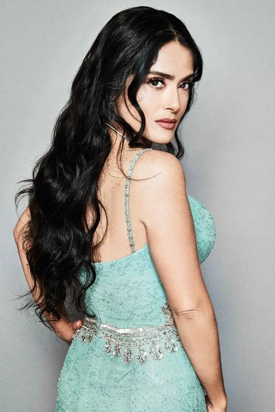 Salma Hayek poses in a light blue dress, with long dark hair, looking over her shoulder in a portrait against a gray backdrop.