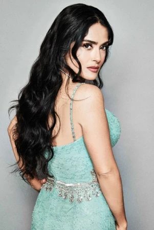 Salma Hayek poses in a light blue dress, with long dark hair, looking over her shoulder in a portrait against a gray backdrop.