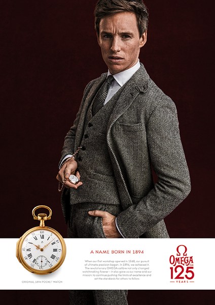 Omega brand advertisement featuring adult male model Eddie Redmayne in grey suit with pocket watch, set against a dark brown background.