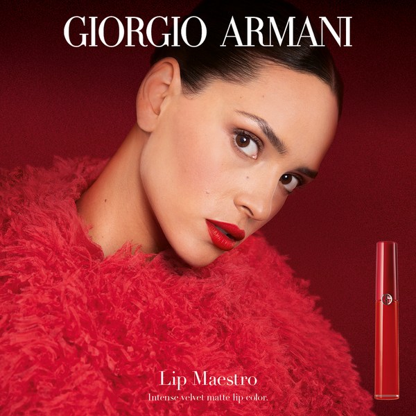 Close-up of a woman with bright red lipstick wearing a fuzzy red jacket, with Giorgio Armani Lip Maestro product against a red background.