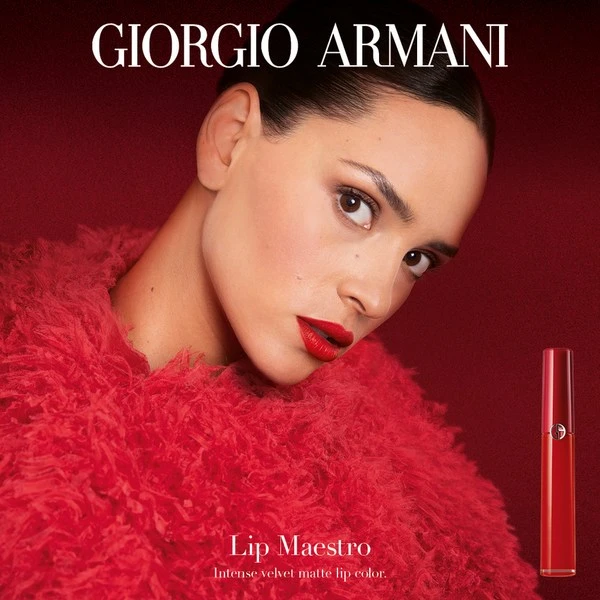 Close-up of a woman with bright red lipstick wearing a fuzzy red jacket, with Giorgio Armani Lip Maestro product against a red background.