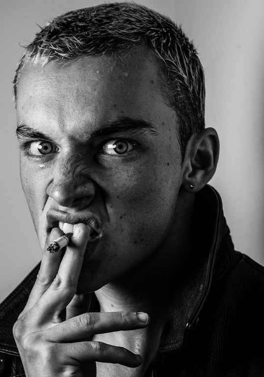 Black and white portrait of a young adult male smoking a cigarette with a scowling facial expression and wearing a jacket.