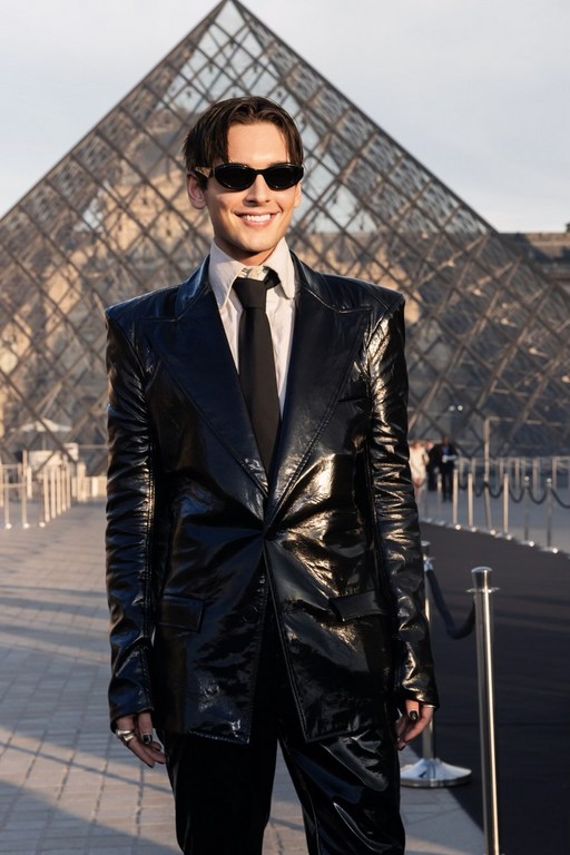 Man poses in a black suit with sunglasses in front of the glass pyramid at the Louvre Museum on a sunny day.