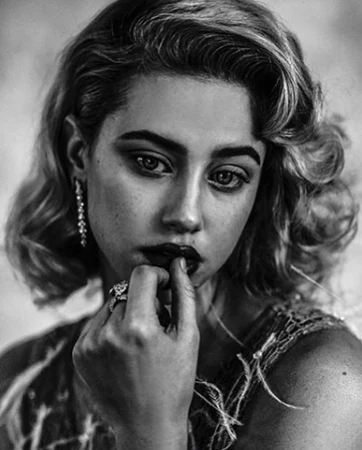Portrait of a woman with wavy blonde hair biting her finger, wearing jeweled earrings and a rings, shot by Damon Baker.