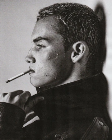 Profile portrait of a young man with a buzz cut and a cigarette leaning against a wall by Damon Baker