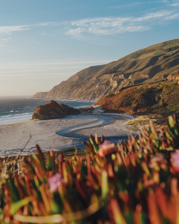 Scenic view of a sandy beach meeting the ocean beneath a grassy hillside with a blue sky and scattered clouds above, capturing coastal beauty.