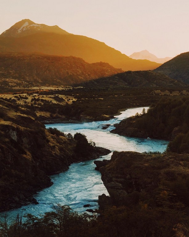 Scenic vista featuring a turquoise river snaking through a valley, flanked by hills and distant mountains, all bathed in warm light.