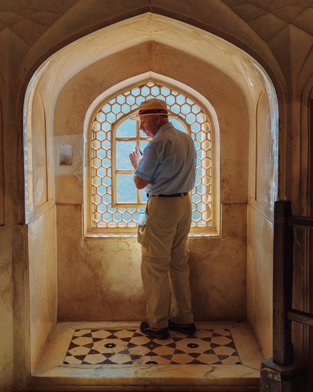 Adult male in hat looks out of an ornate, geometric window in a tan stone interior with patterned marble floor, bathed in soft, natural light.