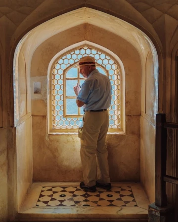 Adult male in hat looks out of an ornate, geometric window in a tan stone interior with patterned marble floor, bathed in soft, natural light.