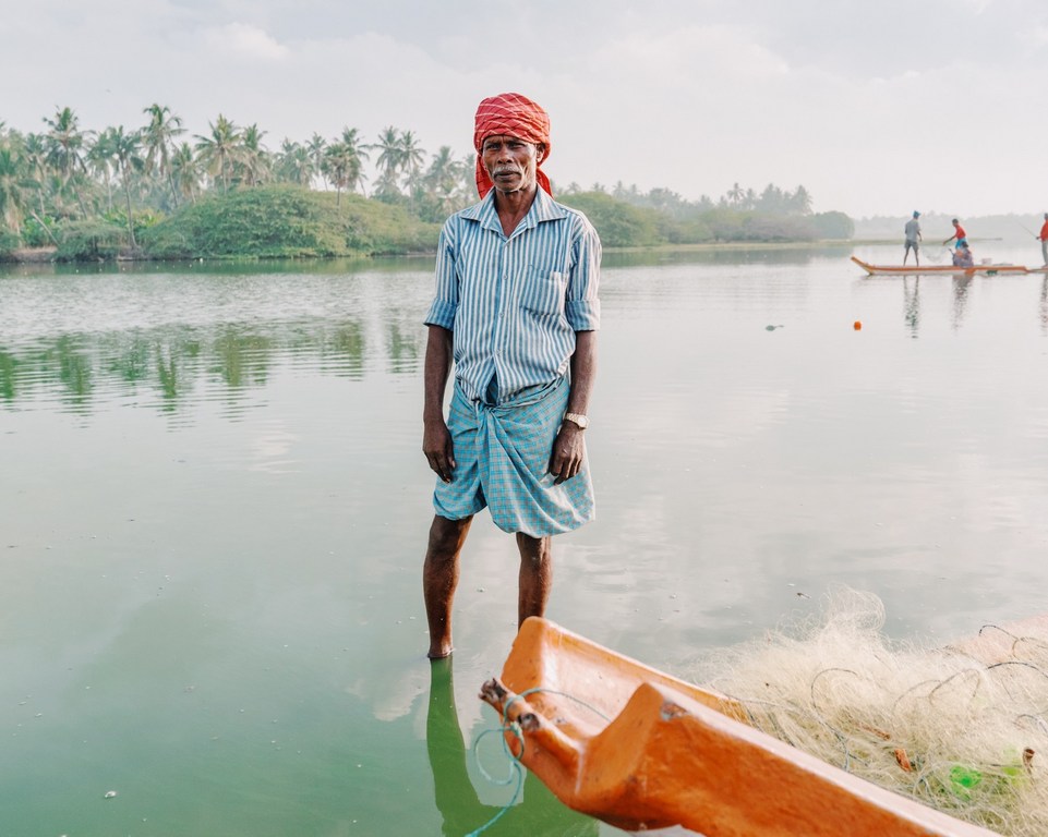 An Indian man stands in shallow water near a boat and fishing net; palm trees line the background on a calm day at the waterside.