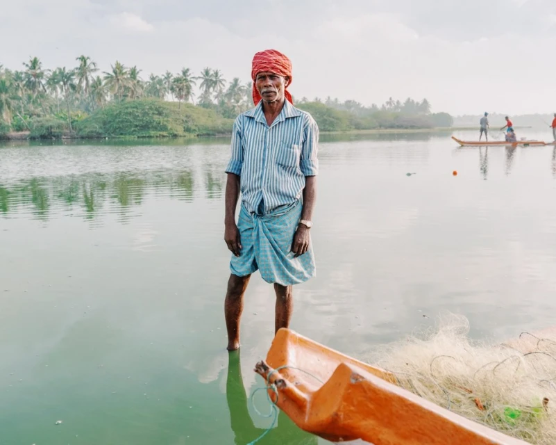 An Indian man stands in shallow water near a boat and fishing net; palm trees line the background on a calm day at the waterside.