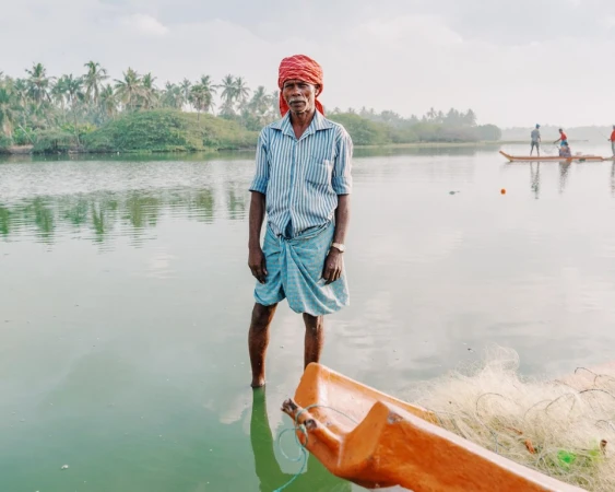 An Indian man stands in shallow water near a boat and fishing net; palm trees line the background on a calm day at the waterside.