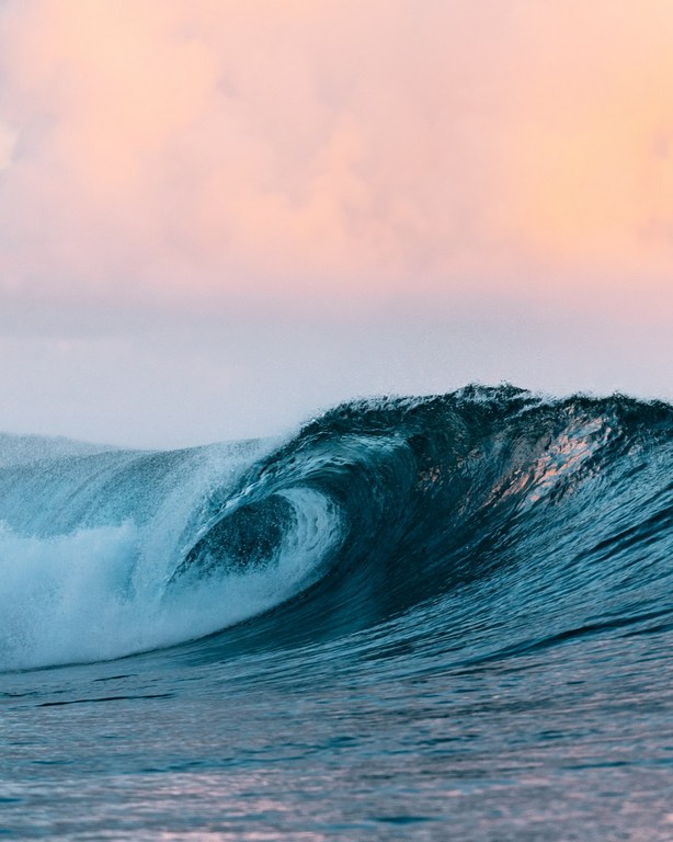 A mesmerizing turquoise wave curls in the ocean against a soft pink and gray sky, showcasing the power and beauty of nature.