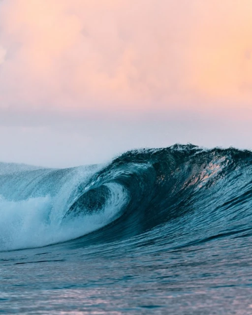 A mesmerizing turquoise wave curls in the ocean against a soft pink and gray sky, showcasing the power and beauty of nature.