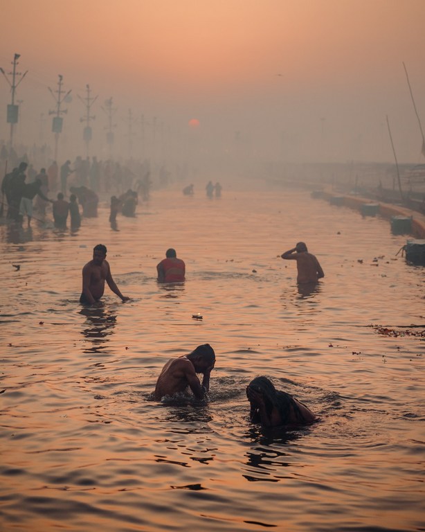 Sunrise view of people bathing in river water; figures in foreground, silhouetted figures and structures in background, orange sky above.