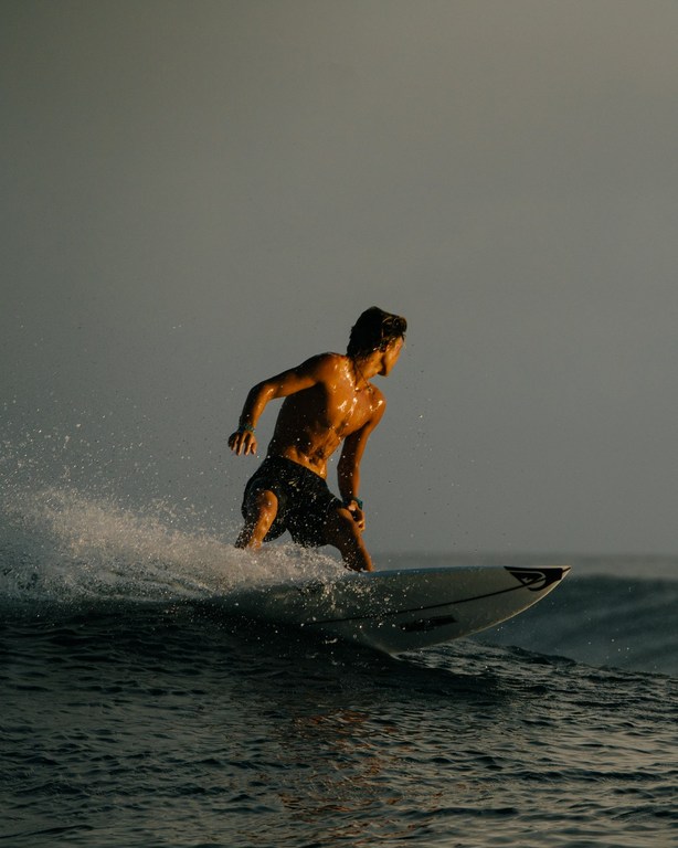 A man surfing on a wave. He is shirtless and wearing dark shorts. The water is dark and the sky is gray, with golden light.