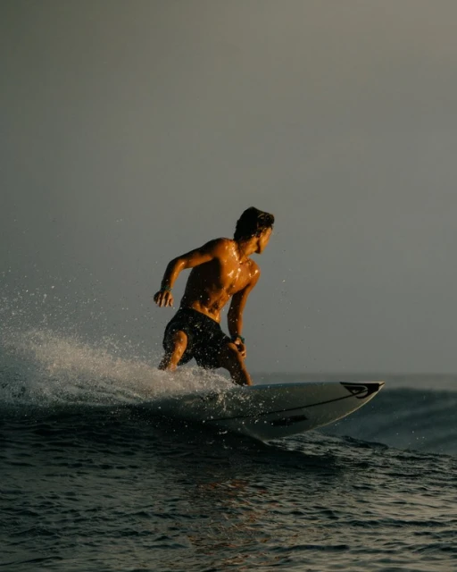 A man surfing on a wave. He is shirtless and wearing dark shorts. The water is dark and the sky is gray, with golden light.