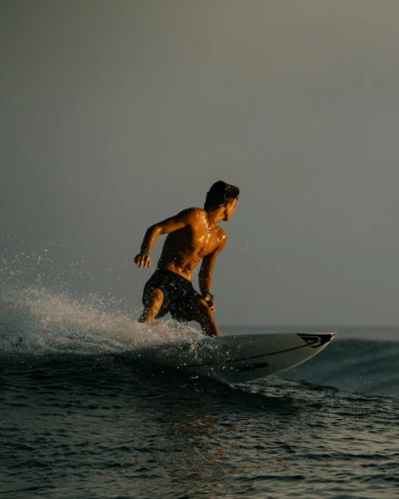 A man surfing on a wave. He is shirtless and wearing dark shorts. The water is dark and the sky is gray, with golden light.