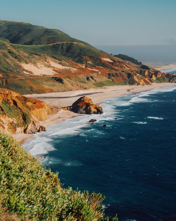 Coastal landscape with rolling hills meeting the ocean, featuring a sandy beach, clear blue sky, and lush green vegetation on cliffs.