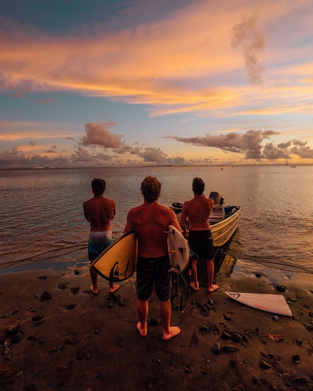 Three surfers stand on a sandy beach near a boat at sunset, holding surfboards, as the sky fills with vibrant orange and pink hues.