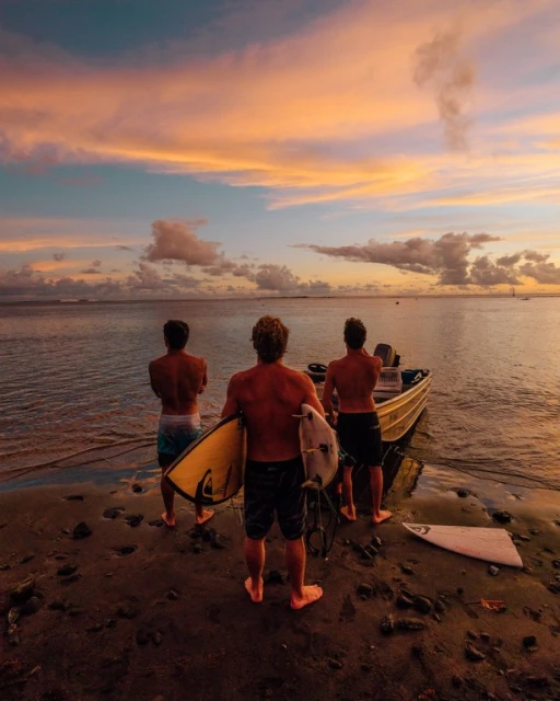 Three surfers stand on a sandy beach near a boat at sunset, holding surfboards, as the sky fills with vibrant orange and pink hues.