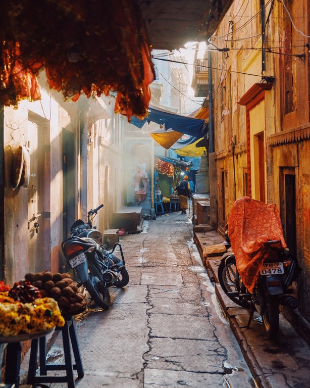 Narrow outdoor alleyway with motorcycles and people in distance, bathed in warm light with building awnings and detailed stone work.