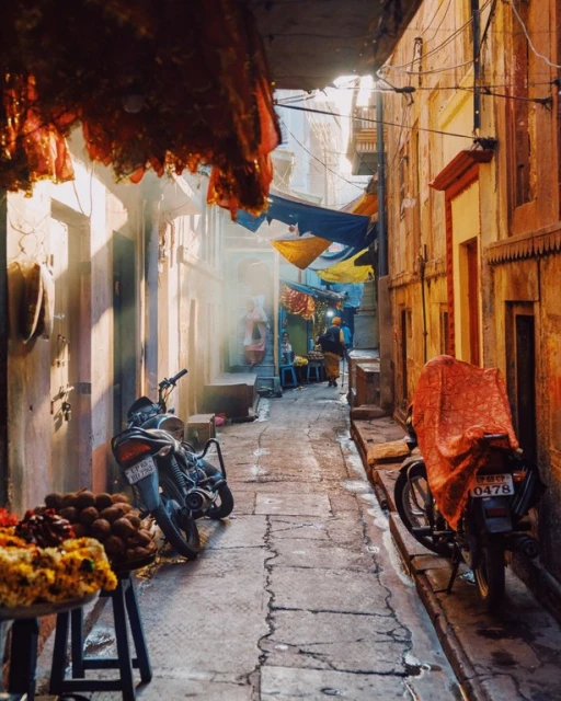 Narrow outdoor alleyway with motorcycles and people in distance, bathed in warm light with building awnings and detailed stone work.