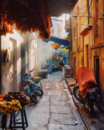 Narrow outdoor alleyway with motorcycles and people in distance, bathed in warm light with building awnings and detailed stone work.