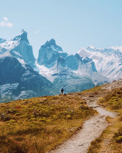 A lone hiker traverses a gravel path on a grassy hill, with views of snow-capped mountains against a clear, bright blue sky.