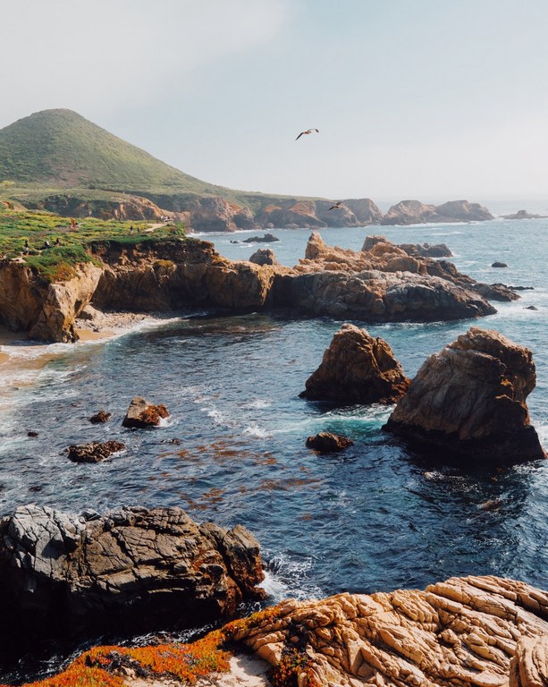 Coastal landscape featuring rocky cliffs, ocean waves, and a distant mountain with green vegetation under a clear sky. 